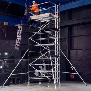 BoSS 3t ladderspan scaffold tower Worker in high-visibility jacket and hard hat operating on the top platform of a tall BoSS Ladderspan 3T mobile aluminium scaffold tower, featuring integral ladders, stabilisers, braced frames, and locking castors in an indoor setting