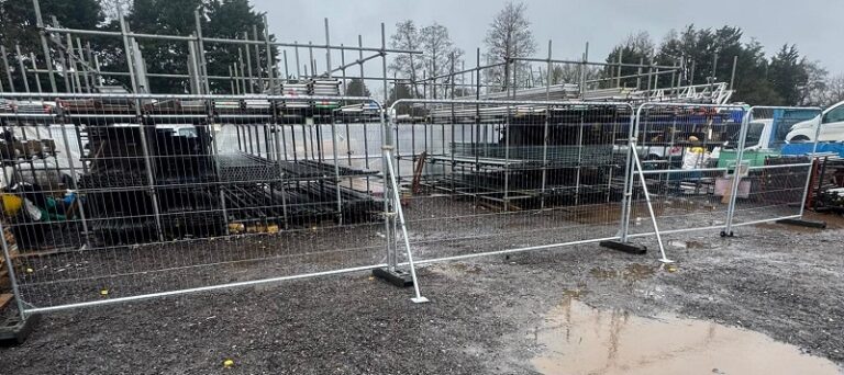 Installed Heras temporary fencing system with galvanised mesh panels and diagonal stabiliser braces, featuring weighted base feet for stability on a construction site yard
