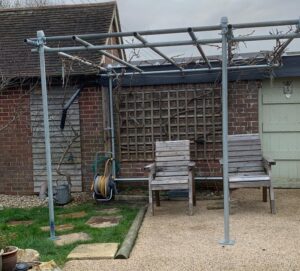 Scaffold tube pergola built from used 10ft tubes and double couplers, supporting trained grapevines over a gravel seating area in a Kent garden