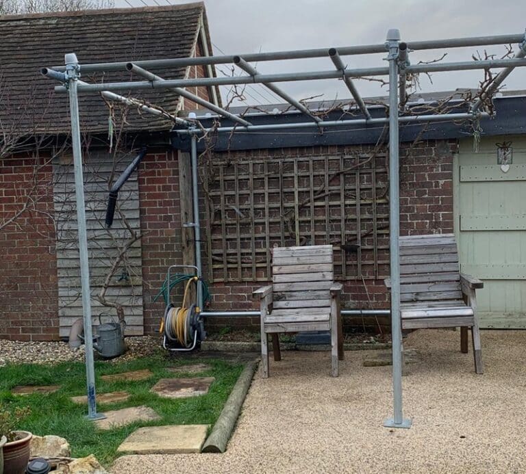 Scaffold tube pergola built from used 10ft tubes and double couplers, supporting trained grapevines over a gravel seating area in a Kent garden